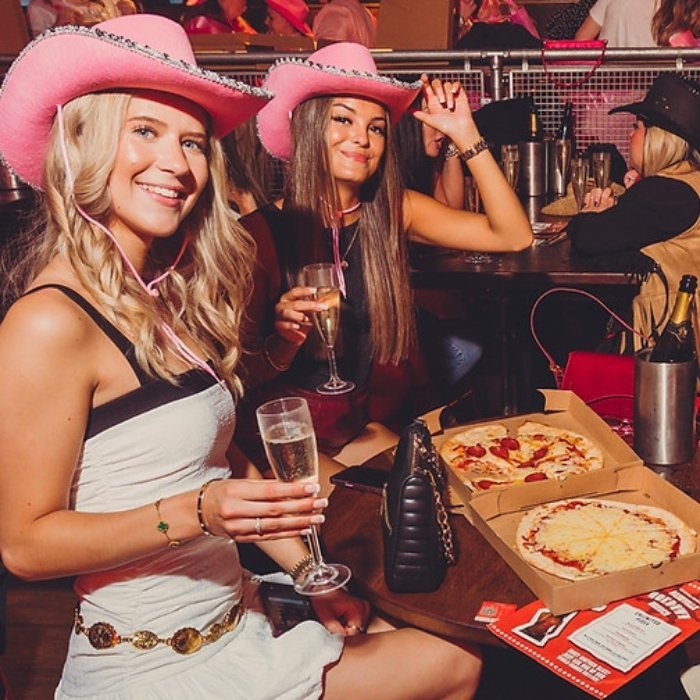 Friends in pink cowboy hats enjoying pizza and prosecco at Coyote Ugly